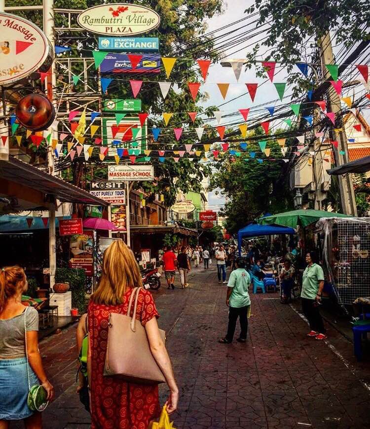 Soi Rambuttri street in Bangkok with colorful flags, market stalls, and pedestrians walking