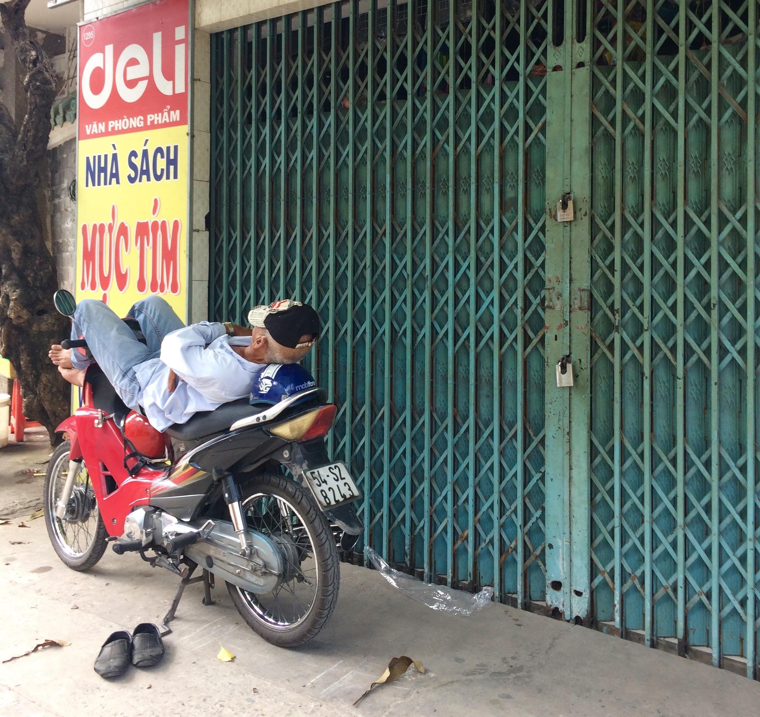Person resting on a motorbike beside a closed shop in Thailand